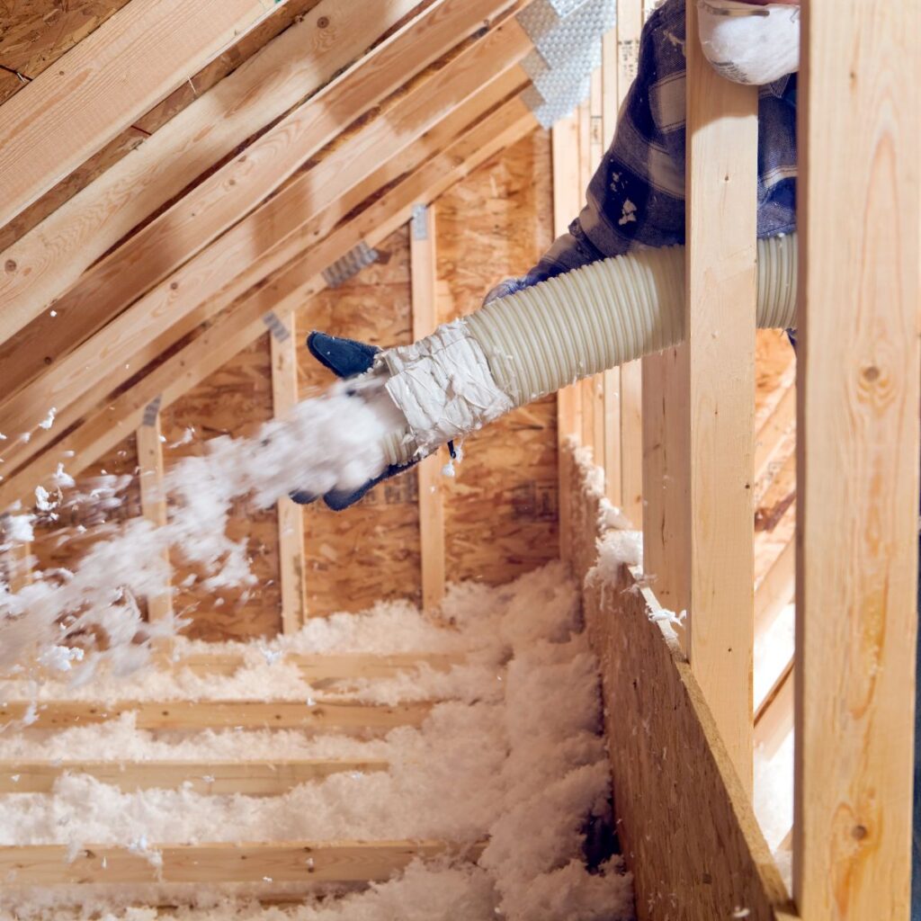 a large white plastic hose blowing small, fluffy pieces of white insulation into an unfinished attic space