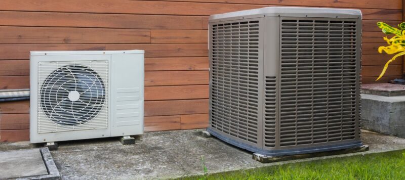 a gray heat pump installed next to a white ductless unit outside a home with wood siding